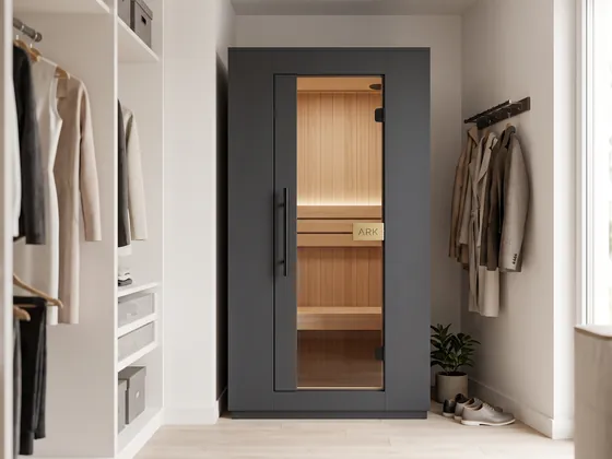 ARK Sauna Compact in black: a one-person cabin with a glass door and a gold ARK plaque, standing in a walk-in closet